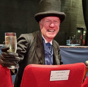 A man in a top hat and gloves sits in a theatre seat, smiling and holding a glass of champagne. The seat has a plaque with a dedication.
