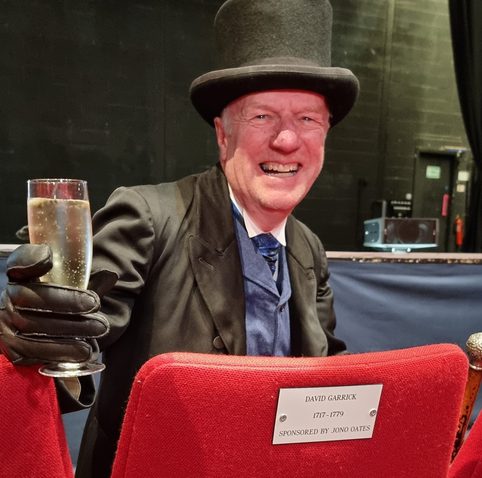A man in a top hat and gloves sits in a theatre seat, smiling and holding a glass of champagne. The seat has a plaque with a dedication.