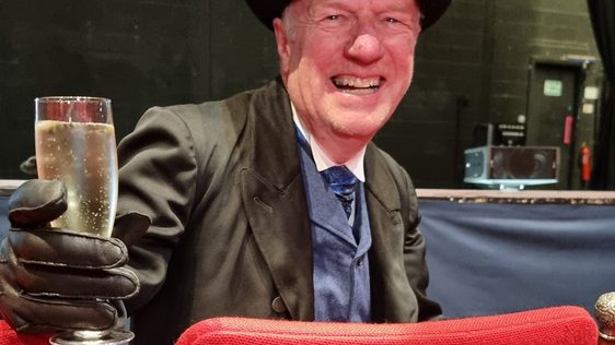A man in a top hat and gloves sits in a theatre seat, smiling and holding a glass of champagne. The seat has a plaque with a dedication.