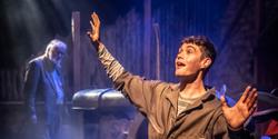 Production shot of Farmboy in the Garrick Studio, a young man sits on a chair at the front of the stage with his arms in the air and an old tractor in the background